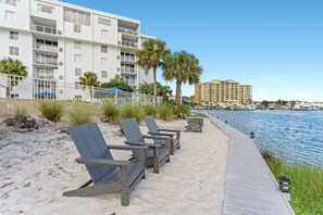 A beachfront promenade with lounge chairs, a white building with balconies, and a body of water with boats. - Dolphin Point 305C🛥️Destin Harbor🌊Waterfront (Destin)