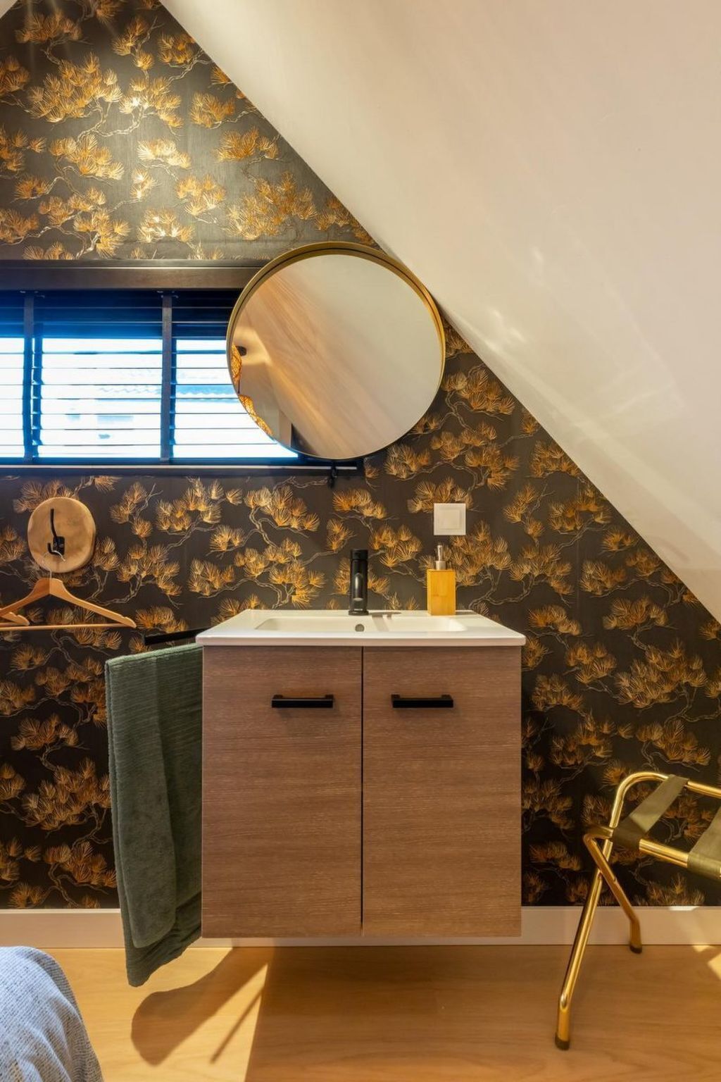 A bathroom with a vanity, a round mirror, a towel, and a window with blinds.