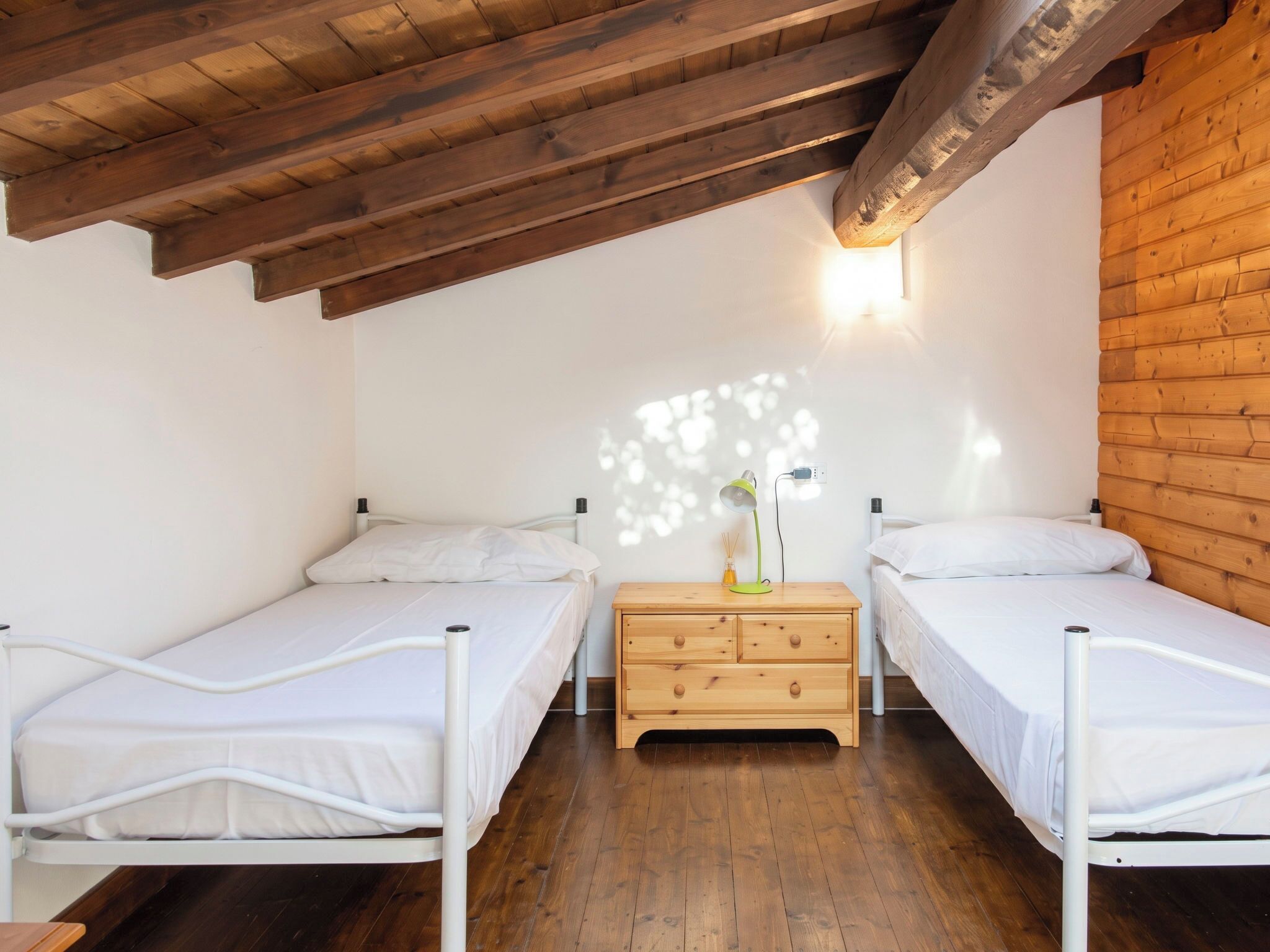 Quarto com duas camas de solteiro, piso e teto de madeira, e luminária fixada na parede.