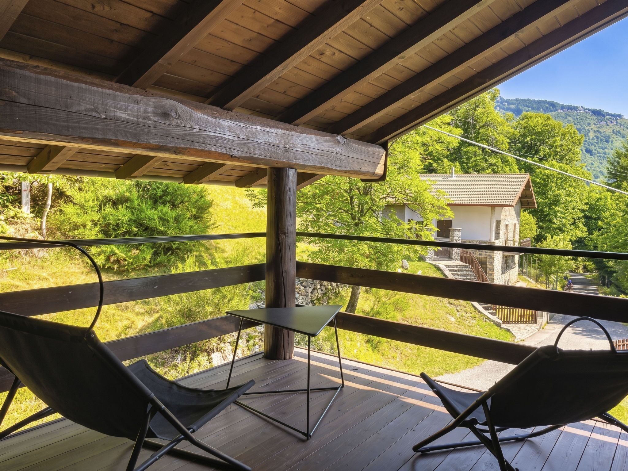 Terraza de madera con dos sillas negras y una mesita, con vistas a un paisaje rural que incluye una casa y árboles.