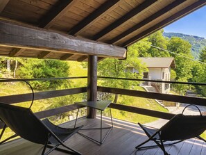 Eine Holzterrasse mit zwei schwarzen Stühlen und einem kleinen Tisch, mit Blick auf eine ländliche Landschaft mit einem Haus und Bäumen.