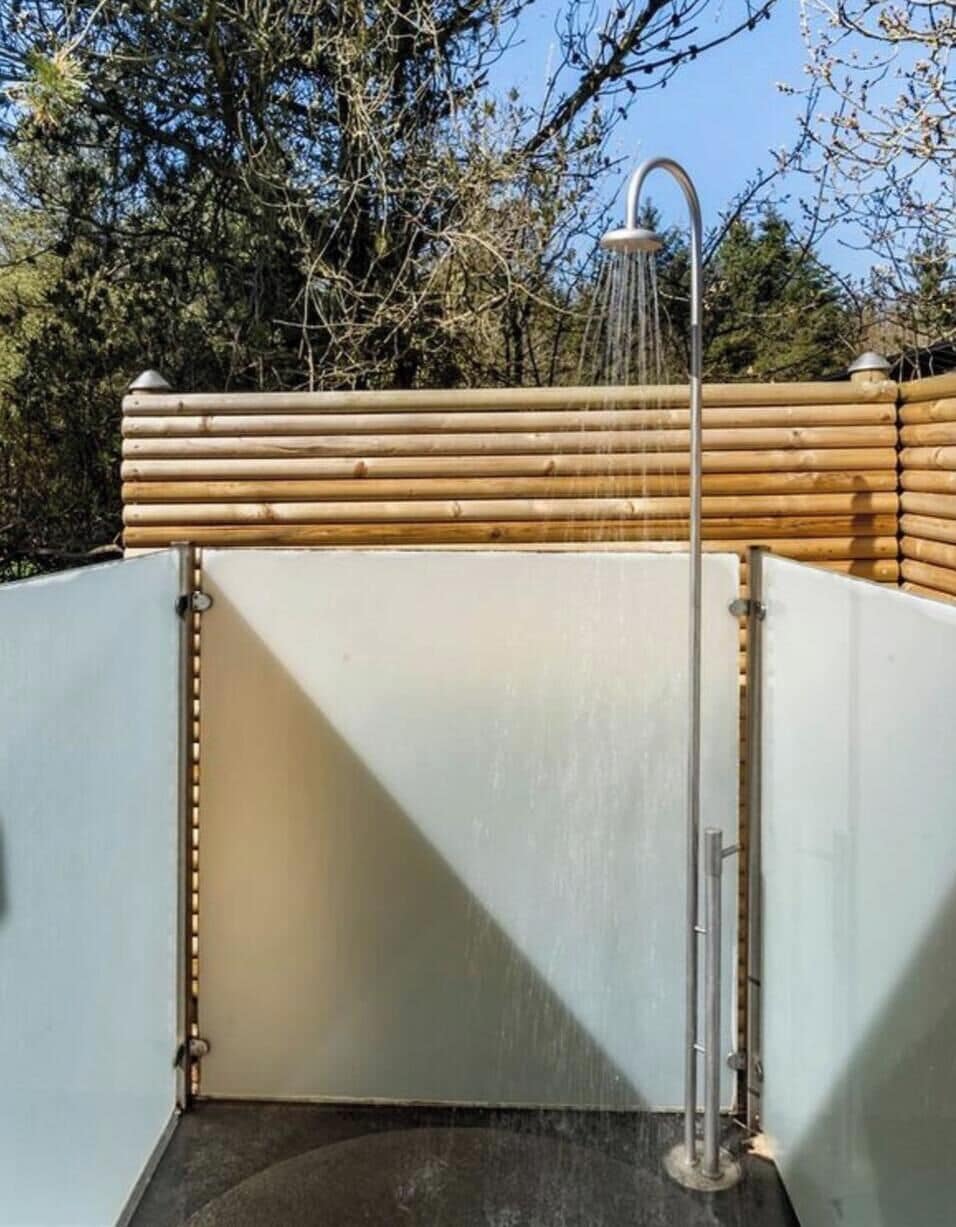 Chuveiro externo com deck de madeira, chuveiro de metal e céu azul ao fundo.