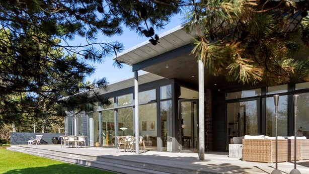 Une maison moderne dotée de grandes fenêtres en verre, d’un patio avec des meubles d’extérieur et d’une pelouse bien entretenue.