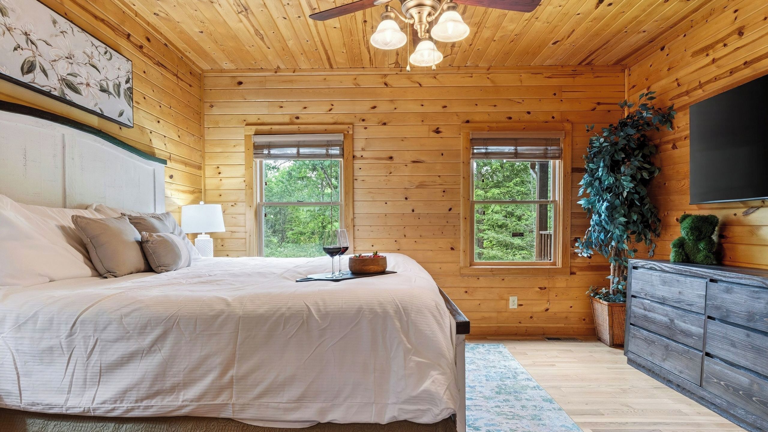 Ein Schlafzimmer einer Holzhütte mit Bett, Nachttisch, Lampe, Fenstern und einem Fernseher.