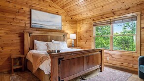 Ein Schlafzimmer einer Holzhütte mit Bett, Nachttisch, Lampe und einem Fenster mit Blick auf Bäume.