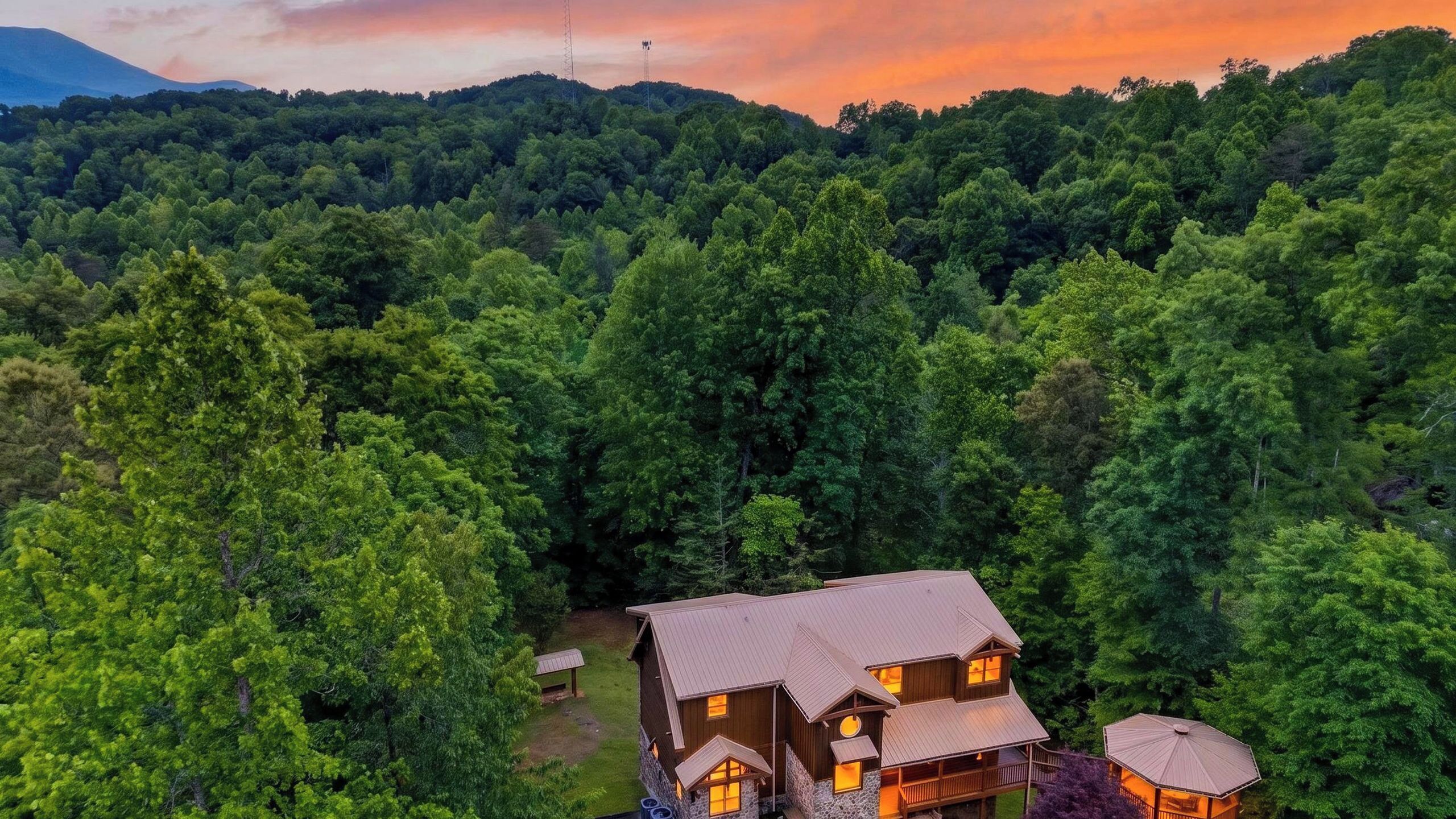 A two-story cabin with a stone foundation and a metal roof, surrounded by dense forest.