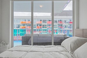 Ein Hotelzimmer mit großem Fenster, einem Bett mit weißer Bettwäsche und Blick auf farbenfrohe Gebäude.