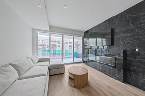 Ein modernes Wohnzimmer mit einer Couch, einem runden Holztisch, einem Flachbildfernseher, der an einer dunklen Marmorwand befestigt ist, und großen, raumhohen Fenstern mit Blick auf eine Stadtlandschaft.