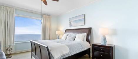 A bedroom with a bed, nightstands, a ceiling fan, a window with curtains, and a view of the ocean.