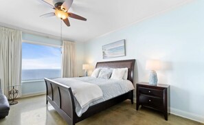 A bedroom with a bed, nightstands, a ceiling fan, a window with curtains, and a view of the ocean. - Indigo West 1703 (Pensacola)