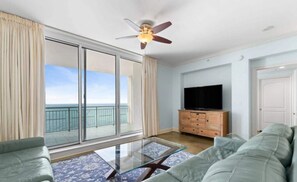A living room with a sofa, a glass coffee table, a TV on a wooden cabinet, and a ceiling fan. - Indigo West 1703 (Pensacola)