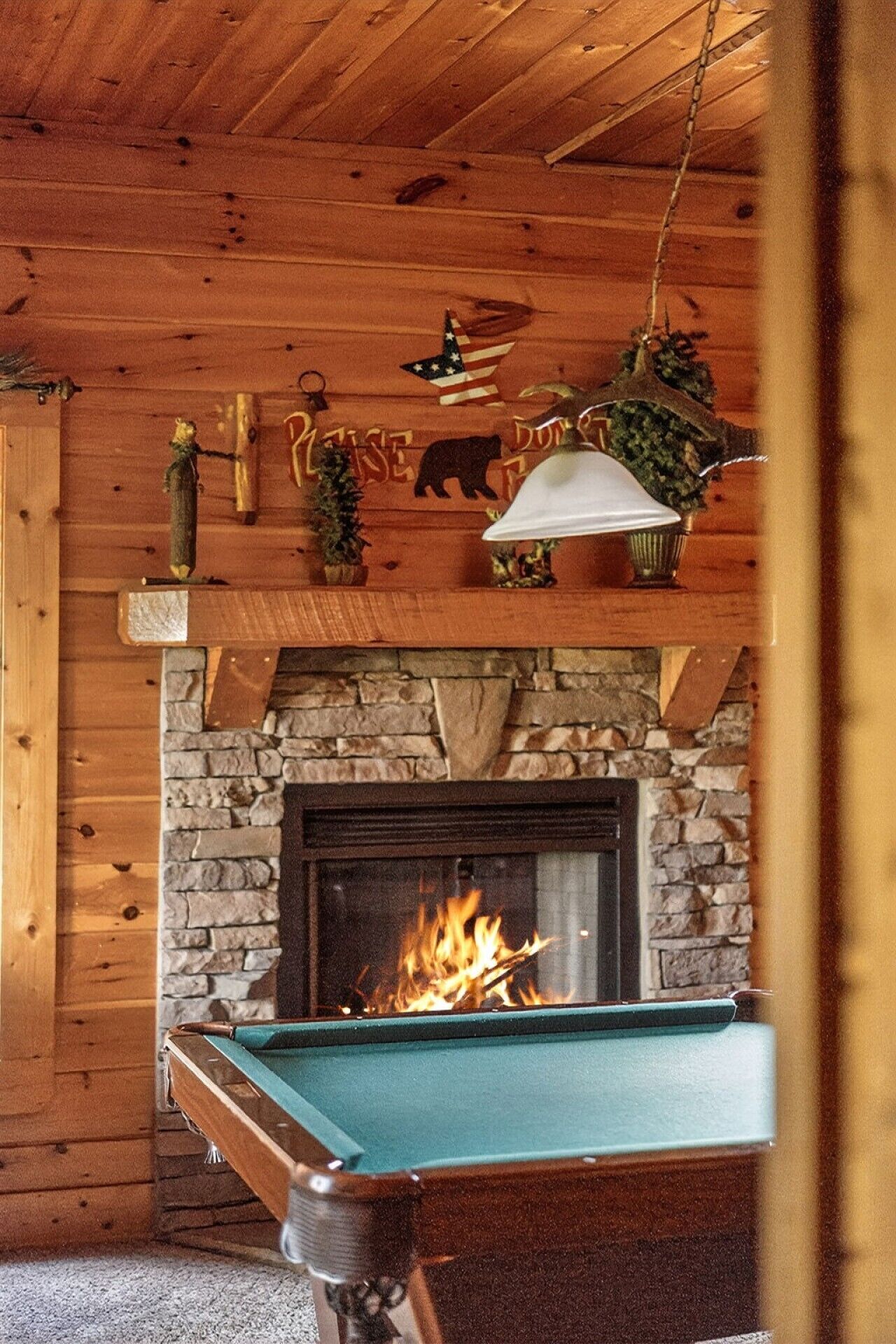 A wooden interior with a stone fireplace, a pool table, and various decorations including a bear, a flag, and a bell.