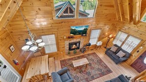 A log cabin interior with a fireplace, a sofa, a dining table, and a chandelier. - Smokies Luxe View: Hot Tub, Fireplaces, Games+MORE (Pigeon Forge)