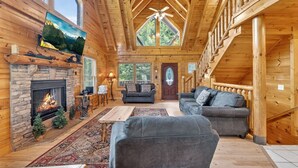 A log cabin interior with a stone fireplace, two sofas, a dining table, and a large window overlooking greenery. - Smokies Luxe View: Hot Tub, Fireplaces, Games+MORE (Pigeon Forge)