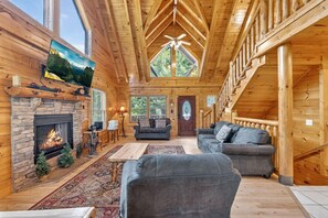 A log cabin interior with a stone fireplace, two sofas, a dining table, and a large window overlooking greenery. - Smokies Luxe View: Hot Tub, Fireplaces, Games+MORE (Pigeon Forge)