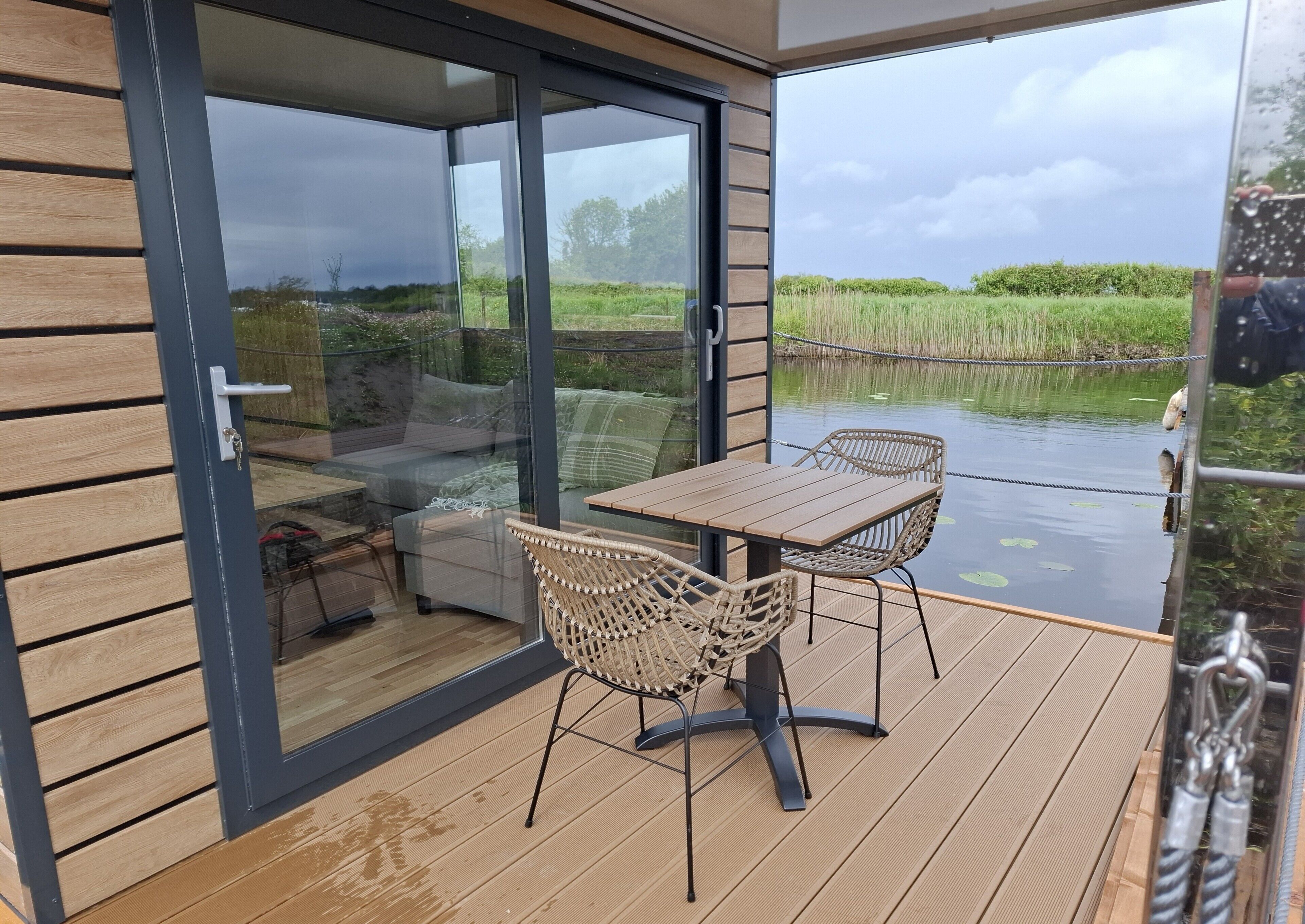 A deck with a table and chairs, a sofa, and a glass door leading to a view of a pond and greenery.. Aquastays offers a unique houseboat experience. Relax by the Water