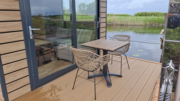 Een terras met een tafel en stoelen, een bank, en een glazen deur die uitkomt op een uitzicht over een vijver en groen.