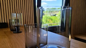 Een houten tafel met twee lege wijnglazen en een glas water, een raam met uitzicht op groen en water, en verticale houten latten aan de muur.