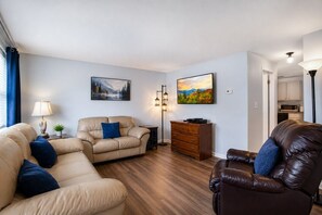 Een woonkamer met een bank, fauteuil en een leren stoel. Er staat een dressoir, een lamp en er hangen twee schilderijen aan de muur.