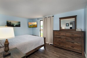 Un dormitorio con cama, cómoda, lámpara, un cuadro y ventana con cortinas.