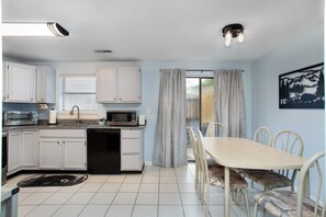 Una cocina compacta con armarios blancos, microondas y lavavajillas. Hay un comedor con mesa y sillas, y una ventana con cortinas.