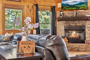 A cozy living room with a leather sofa, a fireplace, a welcome sign, and a window with a view of greenery. - Adohi Lodge | Panoramic Views off the Deck! (Sevierville)