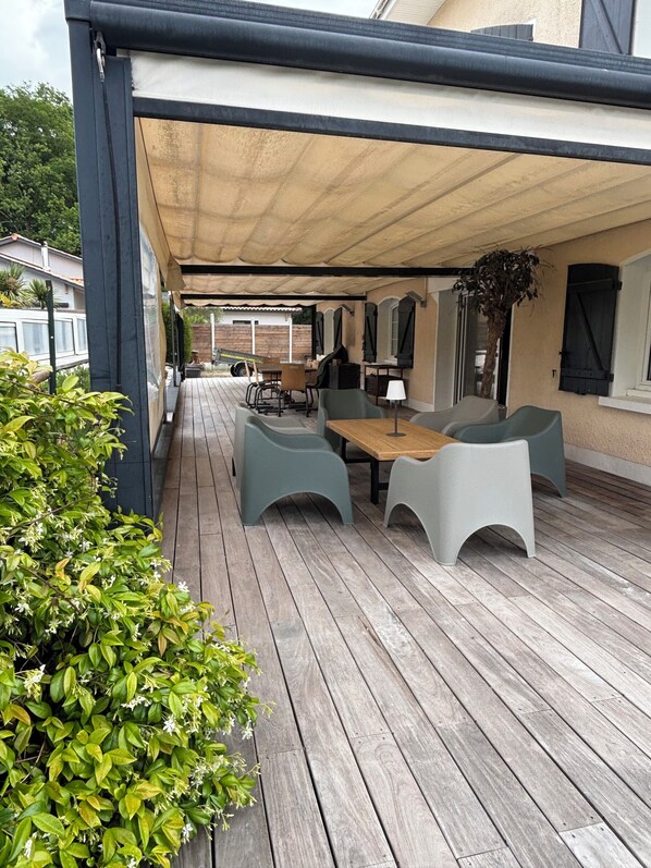 Une terrasse extérieure couverte, avec une table et des chaises en bois, entourée de verdure.