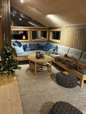 Ein gemütliches Wohnzimmer mit Holzbalkendecke, einem runden Couchtisch, einem grauen Sofa mit blauen Kissen, einem kleinen Weihnachtsbaum und einem Kamin, der durch die Fenster sichtbar ist.