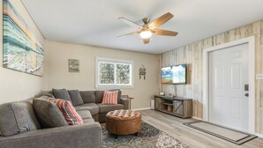 A living room with a sectional sofa, ottoman, ceiling fan, television, and a door. - Peaceful 2-bedroom house with CABANA CLUB in charming Key Colony Beach (Duck Key)