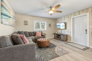 A living room with a sectional sofa, ottoman, ceiling fan, television, and a door. - Peaceful 2-bedroom house with CABANA CLUB in charming Key Colony Beach (Duck Key)