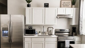 Una cocina moderna con armarios blancos, electrodomésticos de acero inoxidable, un microondas, un lavabo y una encimera con un reloj y un cartel.