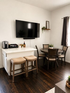 A modern living room with a flat-screen TV, a coffee station, and a dining area with wooden chairs and a round table. - Modern Dallas Escape-FIFA World Cup (Dallas)