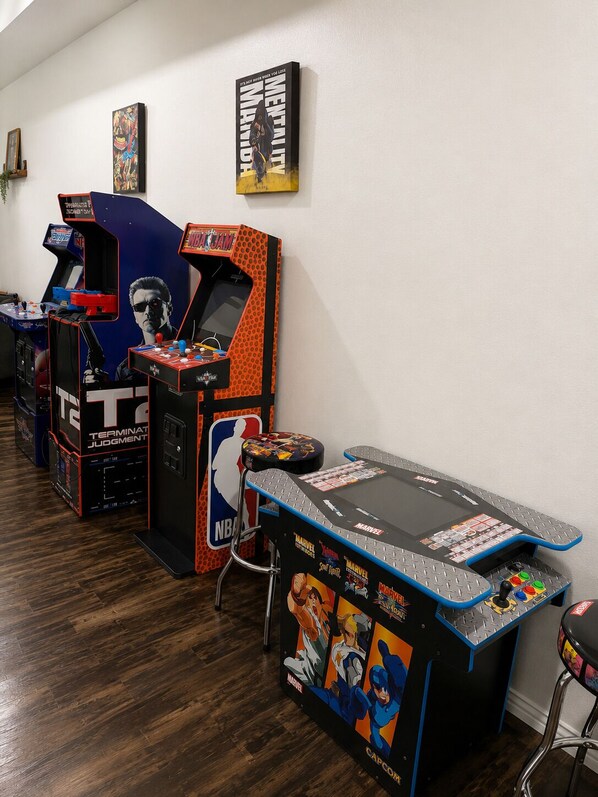 A room with multiple arcade machines, a table with Marvel-themed artwork, and a poster on the wall.