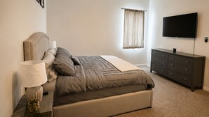 Un dormitorio con cama, mesita de noche, cómoda, ventilador de techo, televisor y ventana con cortinas.