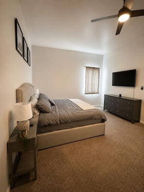 Un dormitorio con cama, mesita de noche, cómoda, ventilador de techo, televisor y ventana con cortinas.