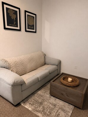 A living room with a grey sofa, a woven throw blanket, a small wooden side table with a candle, and two framed maps of Los Angeles and Dallas on the wall. - Modern Dallas Escape-FIFA World Cup (Dallas)