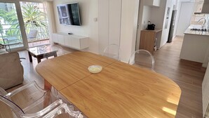 Ein stilvolles Wohnzimmer mit einem Holzesstisch, transparenten Stühlen, einem an der Wand befestigten Fernseher und Blick ins Freie durch große Fenster.