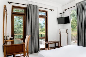 Una habitación de hotel con cama, escritorio, silla, televisión y ventanas grandes con cortinas.