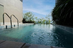 Una piscina con barandillas metálicas, borde de hormigón y vistas a un edificio y vegetación.