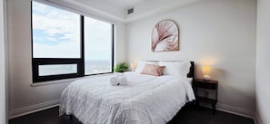 Une chambre avec une grande fenêtre, un lit avec une literie blanche, une table de chevet avec une lampe et une œuvre d’art accrochée au mur.