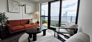 A modern living room with a red sofa, a black coffee table, and a white armchair. There are two framed abstract artworks on the wall, a potted plant, and a view of the city through large windows. - Sky High | Luxe Stay | 2 BR-2 BA | SQ1 | Lake View | Gym | Pool | Parking (Mississauga)