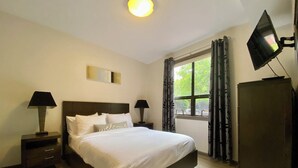 A hotel room with a bed, two bedside tables, a TV, a wardrobe, a window with curtains, and a ceiling light. - Resort-Style Stay in the Heart of Kelowna’s Lower Mission (Kelowna)