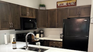 A modern kitchen with dark wood cabinets, a black refrigerator, a white marble countertop, and a 'Welcome to the Lake' sign. - Resort-Style Stay in the Heart of Kelowna’s Lower Mission (Kelowna)