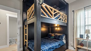 Ein Zimmer mit einem Etagenbett, einer Holzleiter, einem kleinen Regal mit Büchern und einer Lampe sowie einem Fenster mit Vorhängen.