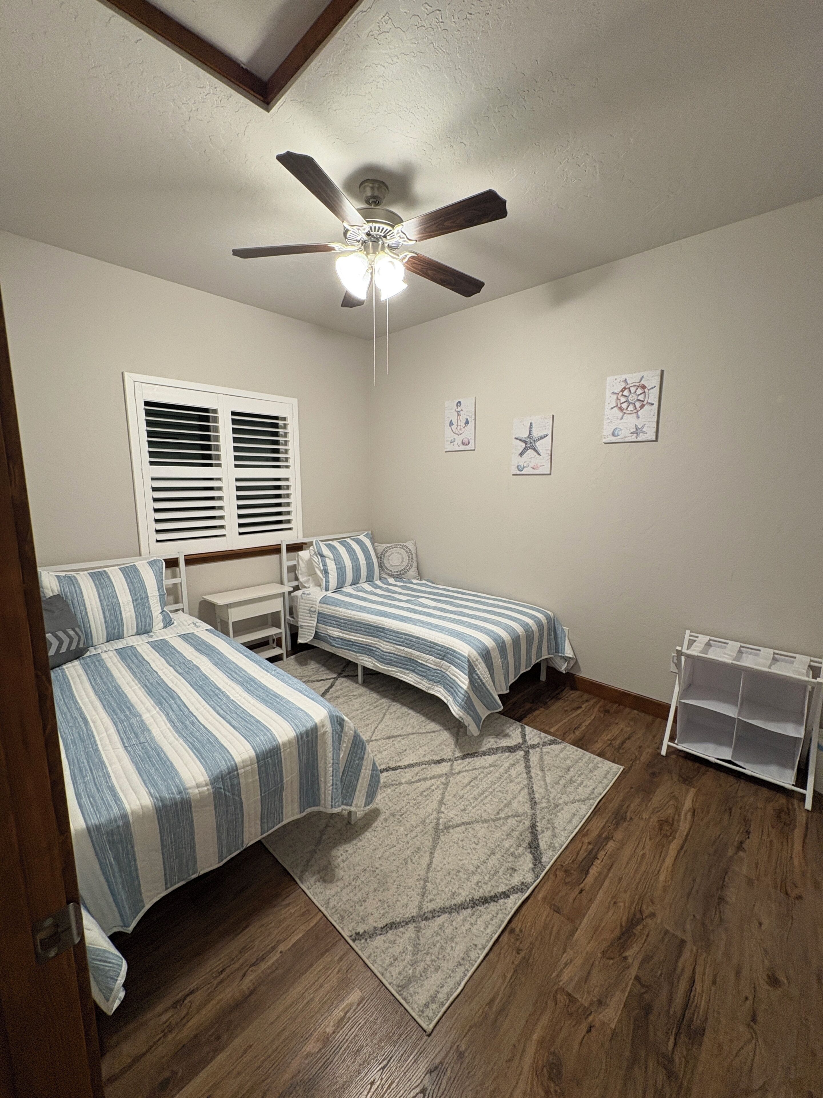 A bedroom with two beds, a ceiling fan, a window with blinds, and wall art.