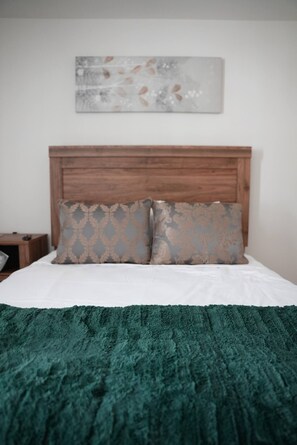 Ein Bett mit hölzernem Kopfteil, zwei Kissen mit Muster, eine grüne, strukturierte Decke und ein an der Wand befestigtes abstraktes Kunstwerk.