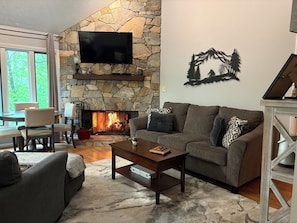 A living room with a stone fireplace, a flat-screen TV, a couch, a coffee table, and a dining area with chairs and a table. - Walk to Slopes, Village & Market. Pool, Dog-Friendly, Bonus Kids Room, Fireplace (Wintergreen Resort)