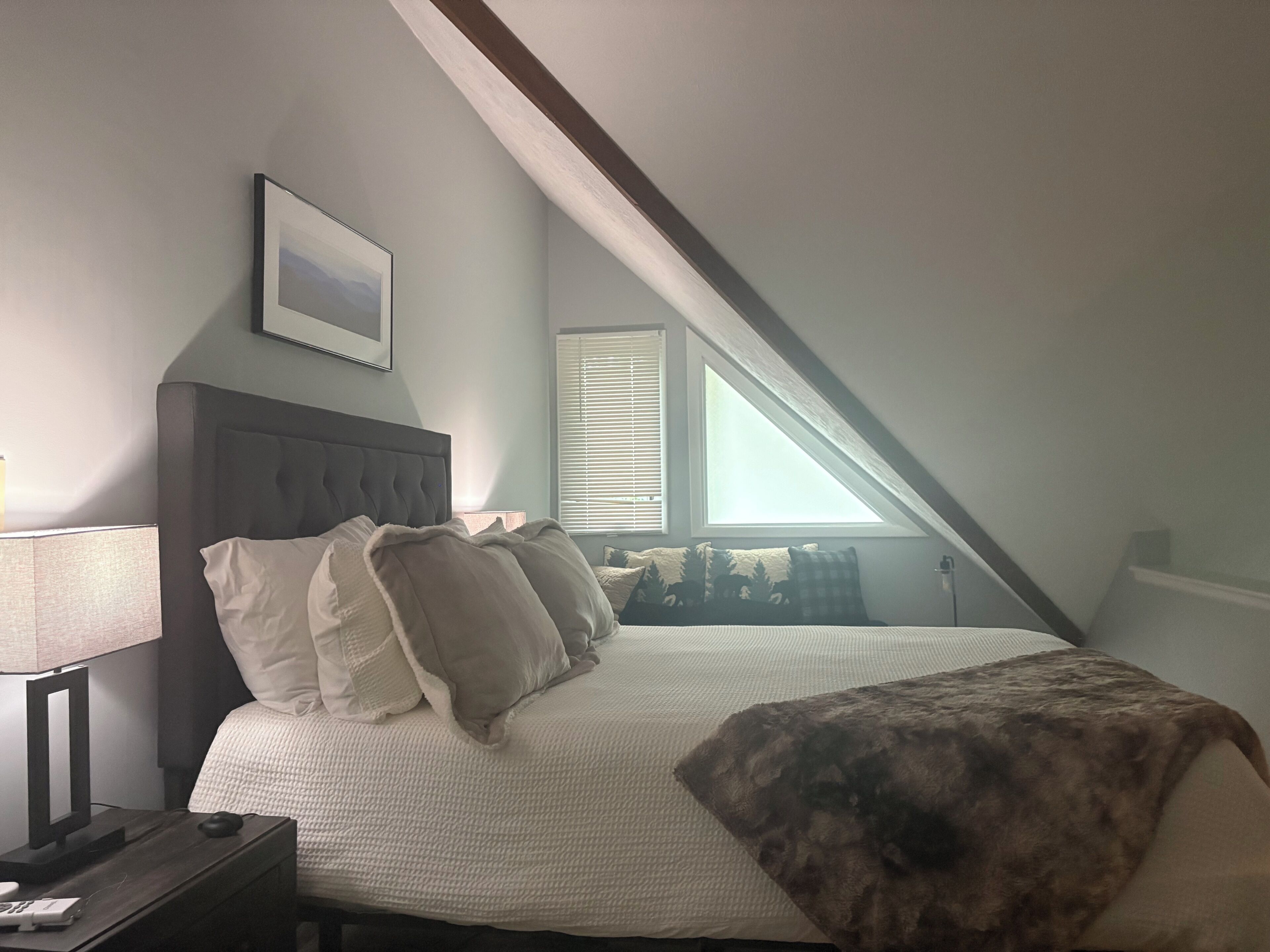 A bedroom with a bed, pillows, a bedside table, a lamp, and a window with blinds.