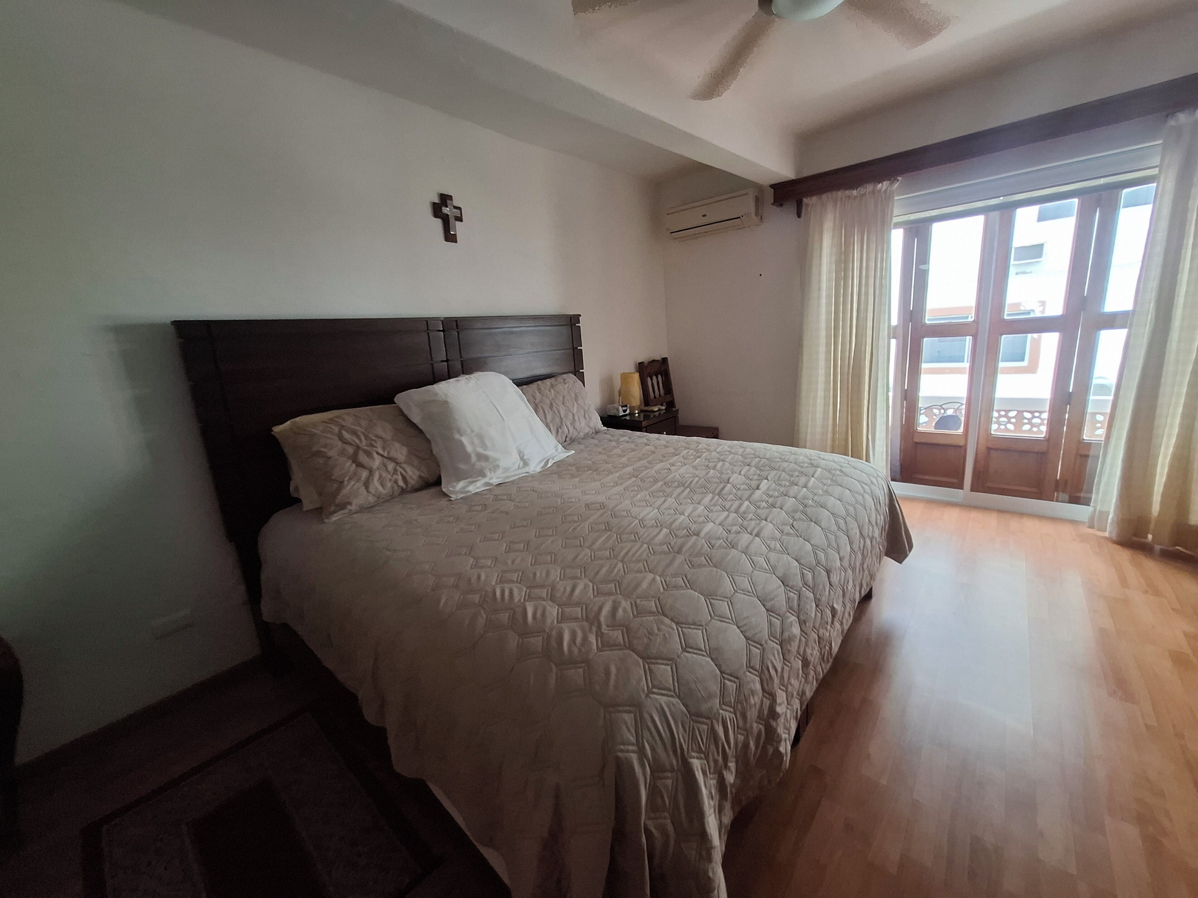 Ein Schlafzimmer mit einem Bett, einem Kreuz an der Wand, einer Klimaanlage und einem fenster mit Vorhängen.