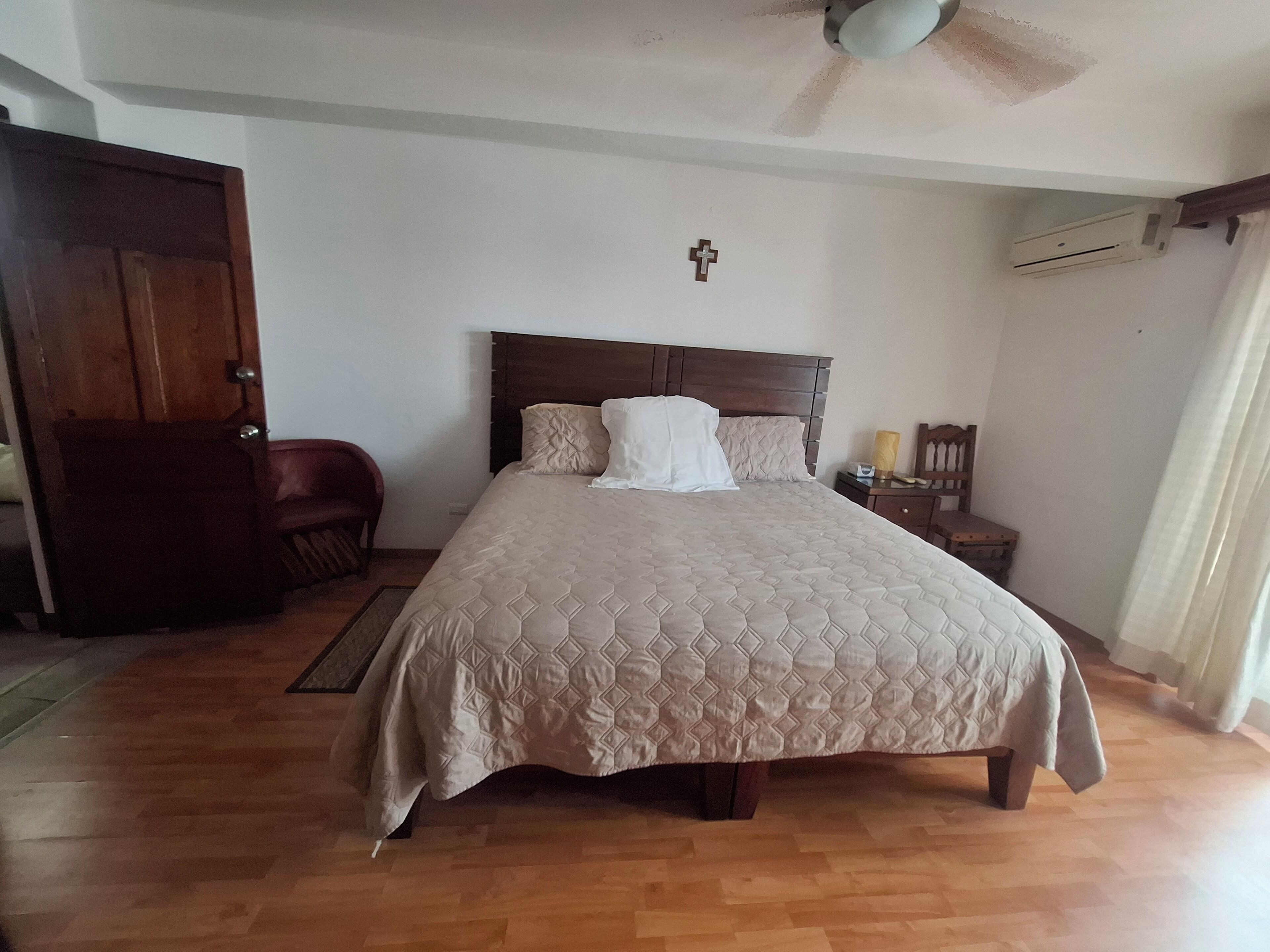 A bedroom with a bed, a wooden headboard, a cross on the wall, an air conditioning unit, a chair, and a nightstand with a lamp.